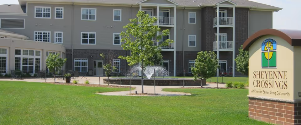Exterior view of Sheyenne Crossings senior living community building with a green lawn, small trees, a water fountain, and a sign displaying the facility name and logo.