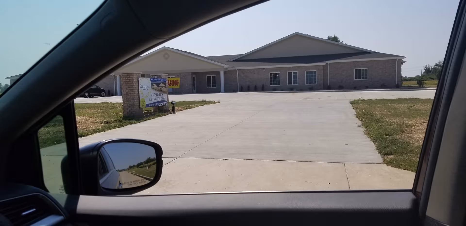 Front exterior view of a memory care facility driveway and signage, seen from a car window.