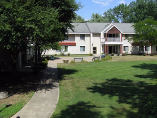 A two-story residential building with balconies and windows, surrounded by a well-maintained lawn and trees under a clear blue sky. A concrete pathway leads towards the building, and there are benches placed on the grass near the building.