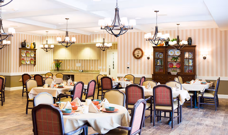 Bright, well-appointed dining room with multiple round tables set for meals, upholstered chairs, chandeliers, and a cabinet displaying plates.