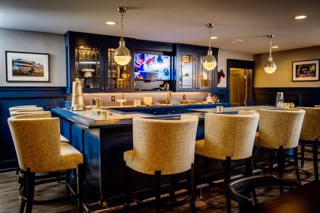 Cozy indoor bar area with a wooden counter, cushioned bar stools, pendant lights, and a television above glass-front cabinets.