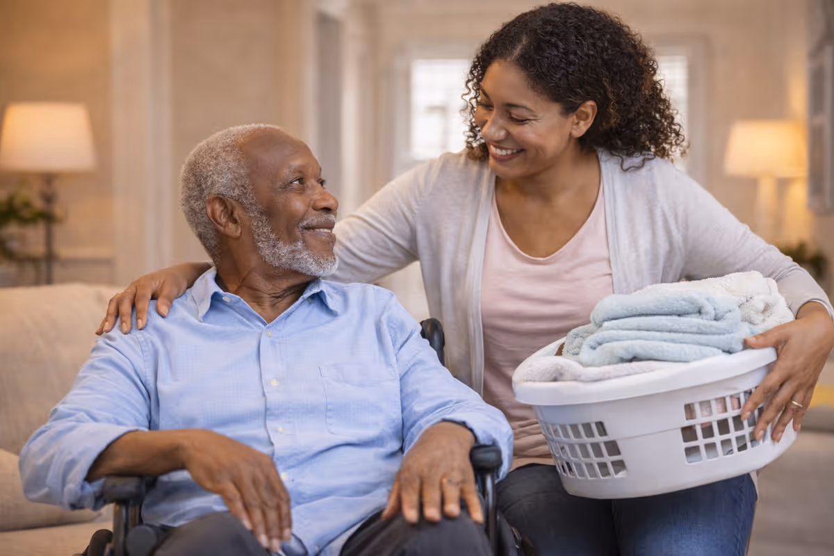 Smiling caregiver assists elderly man in a wheelchair with a laundry basket in a living room.
