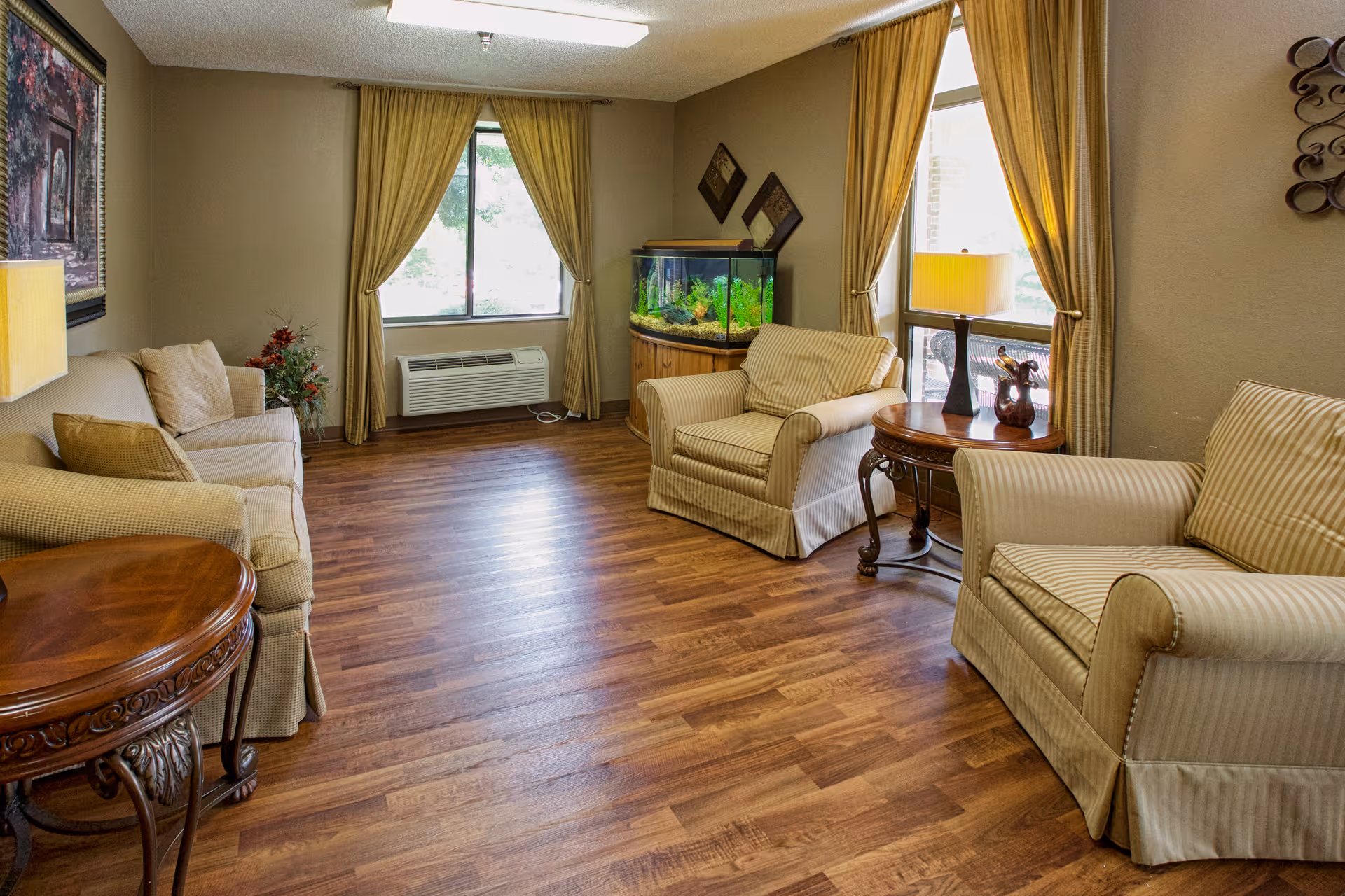 A cozy living room with wooden flooring, beige armchairs and a sofa, two round wooden side tables with lamps, a fish tank in the corner, and windows with beige curtains letting in natural light.