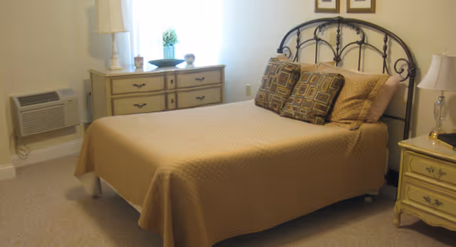 A bedroom with a neatly made bed covered with a beige quilt and three decorative pillows. The bed has a black metal headboard. There is a beige dresser with drawers against the wall, topped with decorative items including a small plant and a lamp. A window with white curtains allows natural light into the room. An air conditioning unit is installed on the wall near the dresser. A nightstand with a lamp and a book is positioned next to the bed.