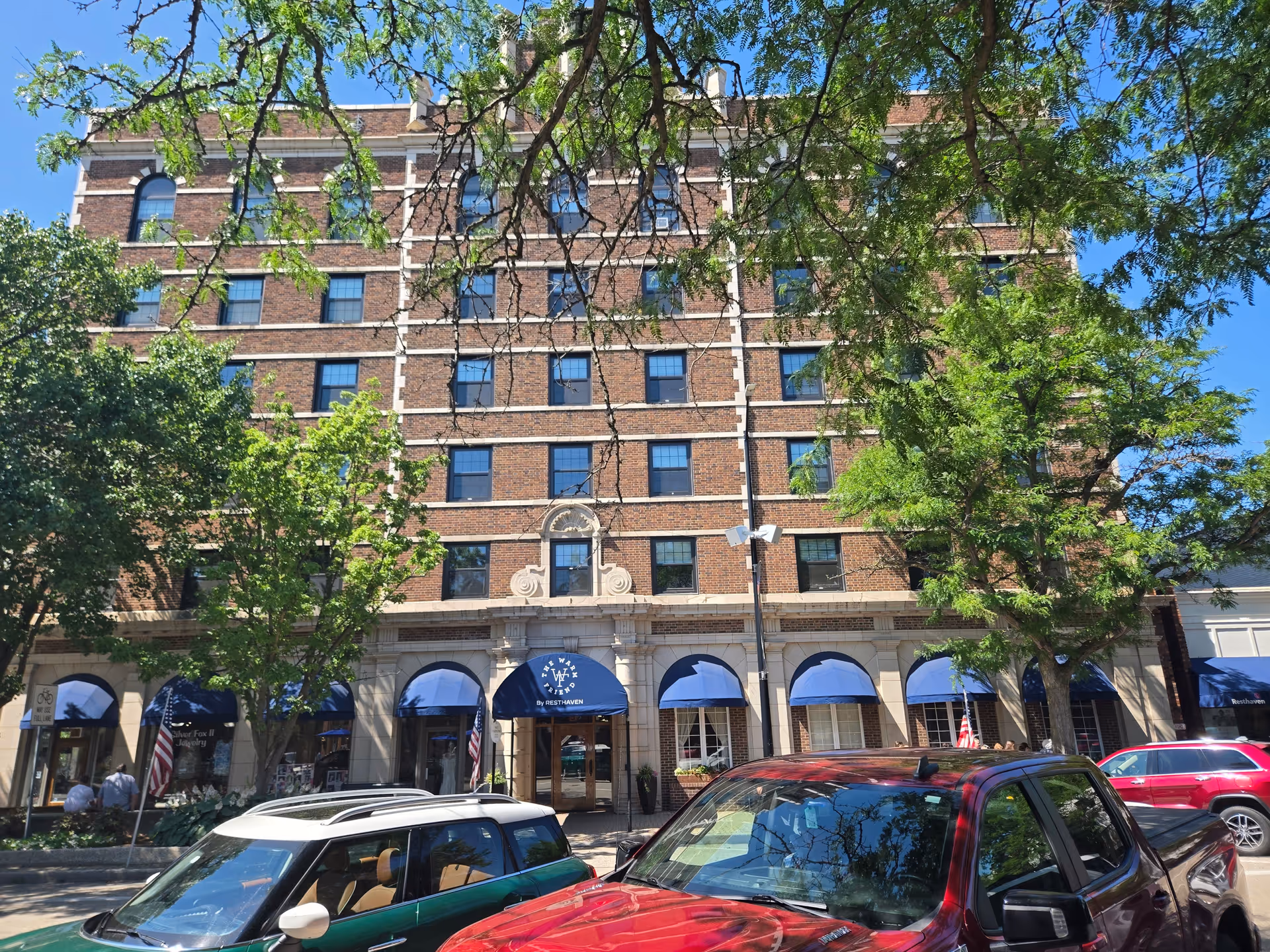Brick senior living building exterior with blue awnings, trees, and parked cars along the street.