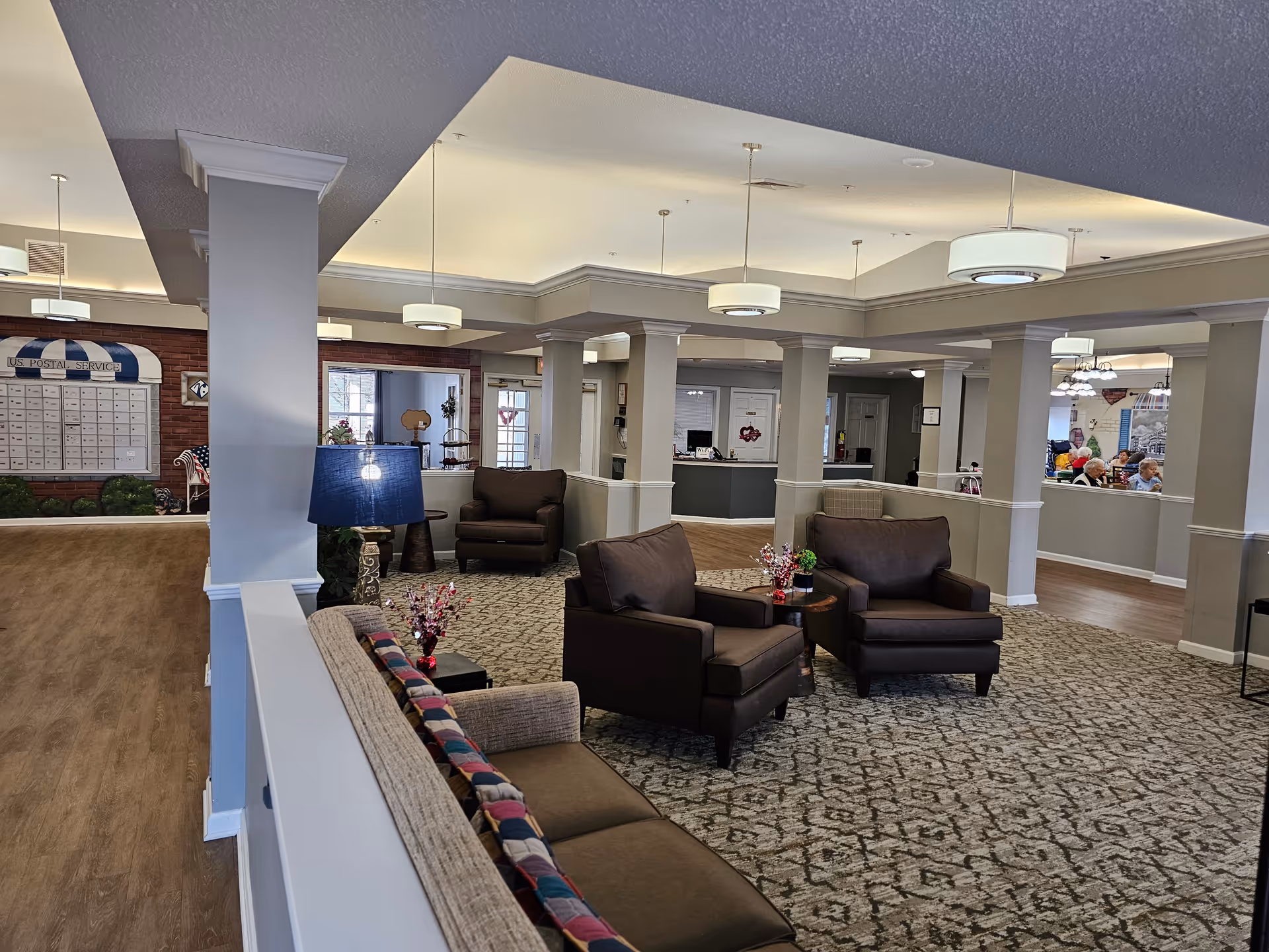 A spacious and well-lit senior living facility common area with comfortable seating including armchairs and a sofa. The room features carpeted flooring, multiple columns, modern ceiling lights, and a US Postal Service mailbox area in the background. There are people visible in a dining area through an open partition on the right side.