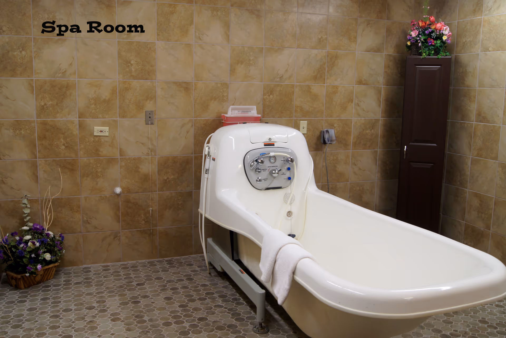 A spa room with a white therapeutic bathtub equipped with control knobs and a towel draped over the side. The room has beige tiled walls and a hexagonal tiled floor. There is a tall dark brown cabinet with a flower arrangement on top and a basket of flowers on the floor.