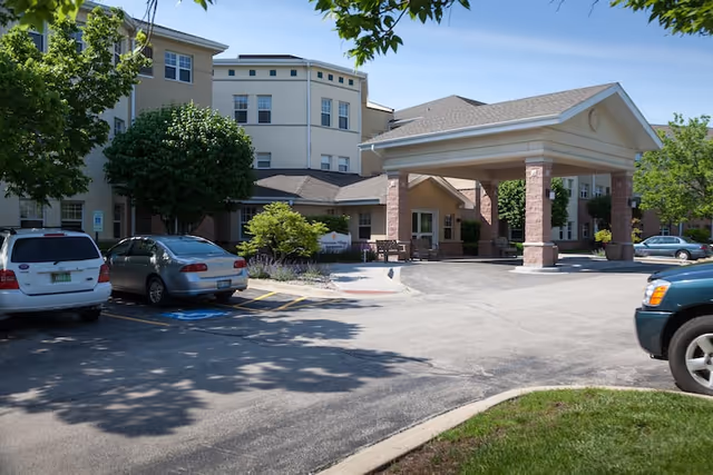 Front entrance of Franciscan Village with a covered porte-cochère, parked cars, and surrounding landscaping.