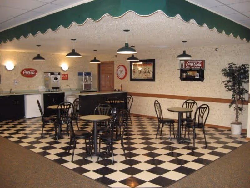 A retro-style dining area with black and white checkered flooring, several round tables with black metal chairs, a counter with beverage machines, Coca-Cola signs on the walls, and a potted plant in the corner.