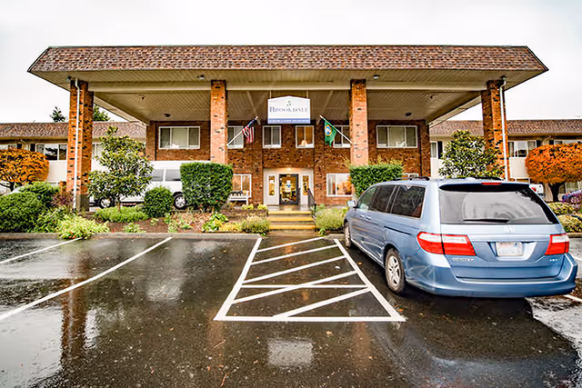Front entrance of a two-story brick Brookdale Federal Way building with a covered portico, flags, shrubs, and a blue minivan in a wet parking lot.