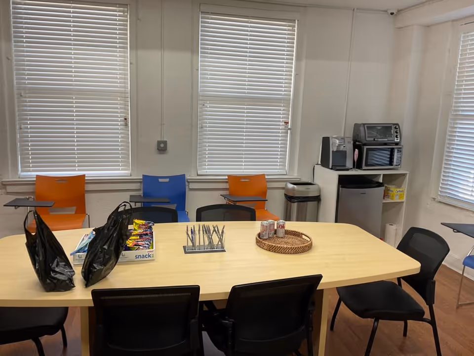 A shared dining room with a long table, colorful chairs, and kitchen appliances.