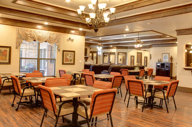 A spacious dining room with multiple square tables and orange cushioned chairs. The room features wooden flooring, beige walls with framed artwork, large windows with decorative valances, and a chandelier hanging from a coffered ceiling with wooden beams. In the background, there is a seating area with dark brown sofas and ceiling fans with lights.