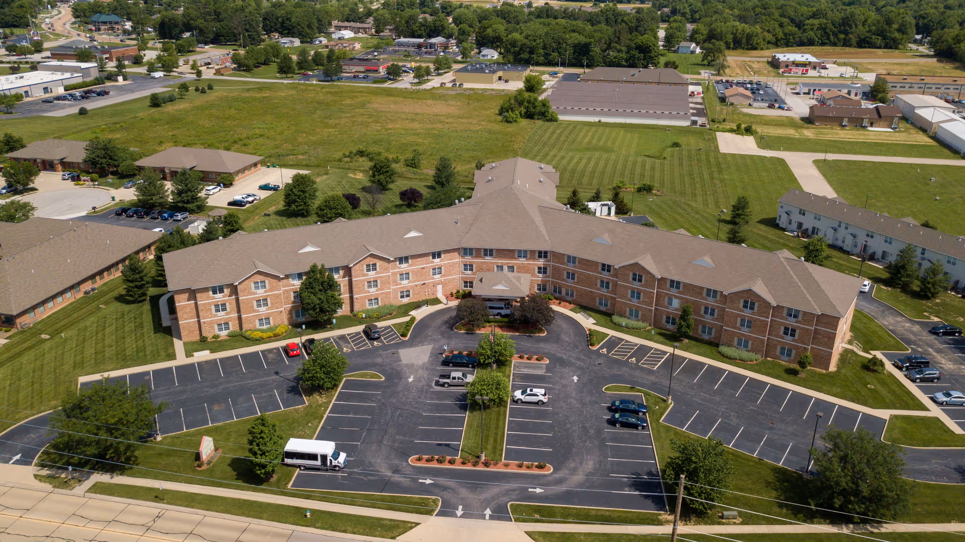 Aerial view of Cambridge House of Ofallon, a three-story brick senior living building with a curved driveway, parking lot, and surrounding lawns.