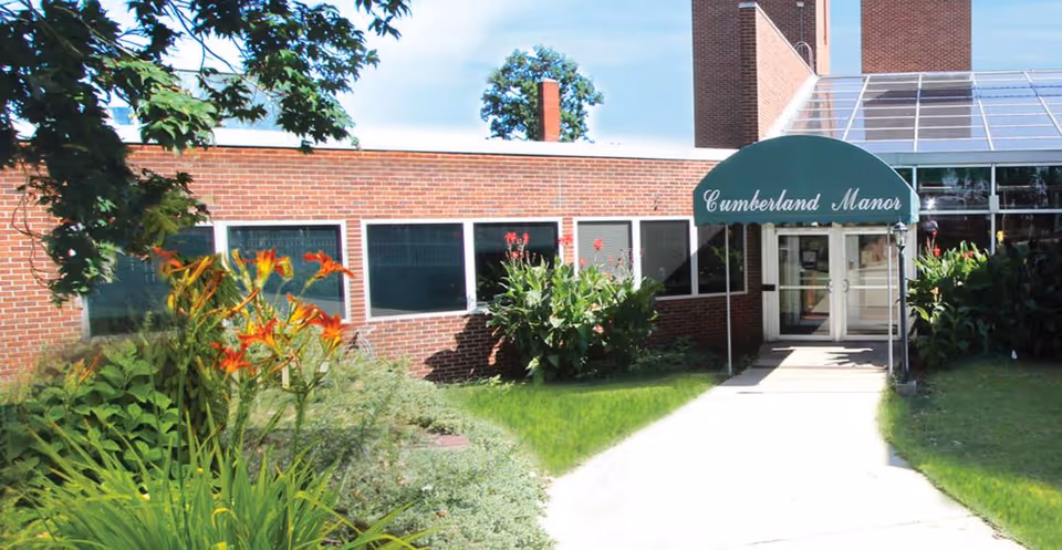 Brick nursing facility entrance with green awning reading Cumberland Manor, surrounded by lush landscaping.