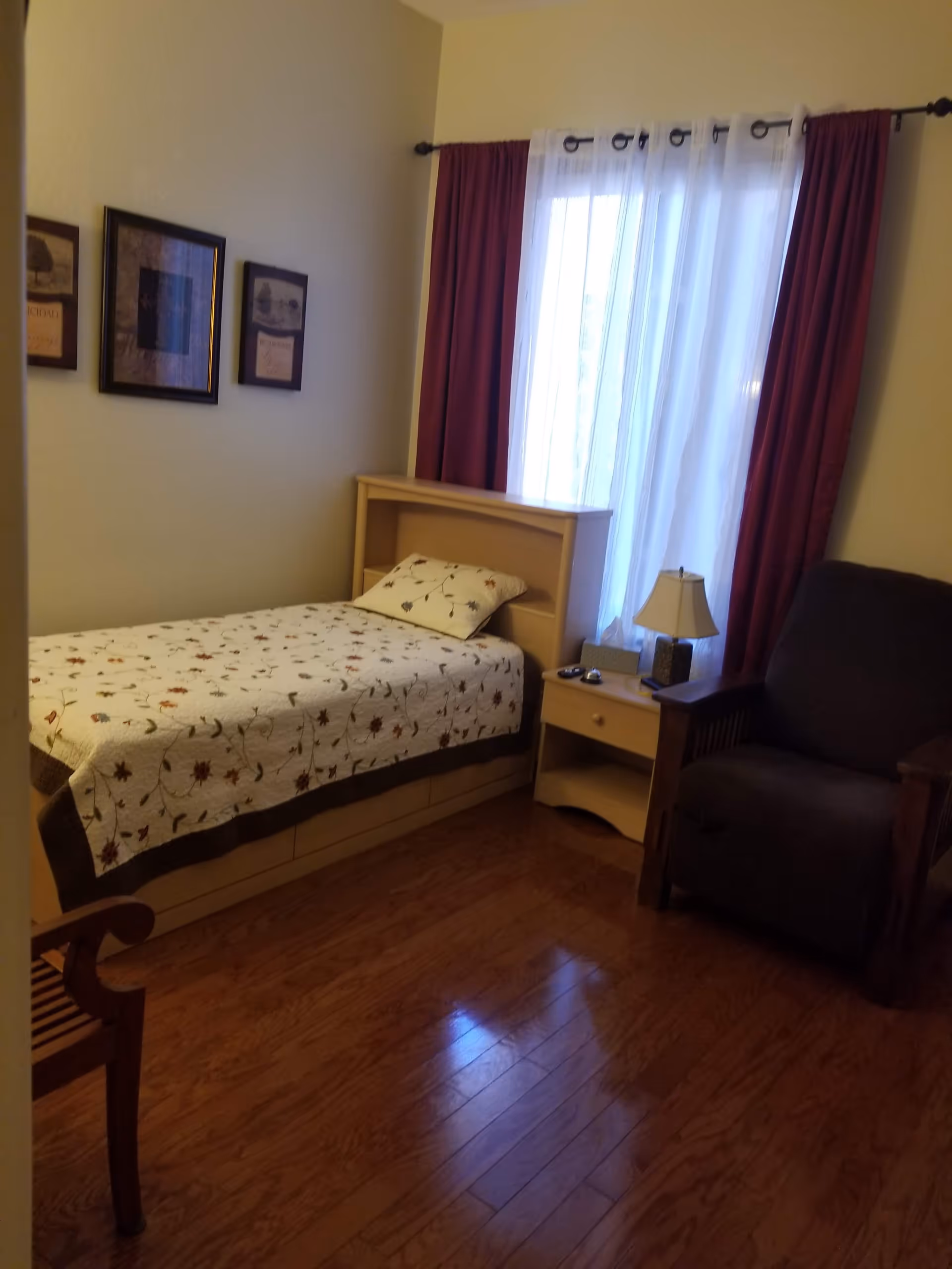 Small furnished bedroom with a single bed, nightstand, lamp and armchair next to a curtained window.