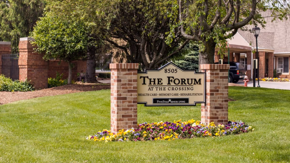Brick-pillared entrance sign reading 'The Forum at the Crossing' on a grassy lawn with flowers, trees, and the facility building in the background.