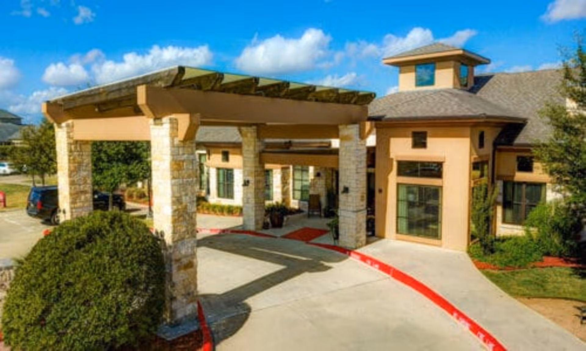 Covered entrance with stone pillars and a driveway leading to a tan senior living facility under a blue sky.
