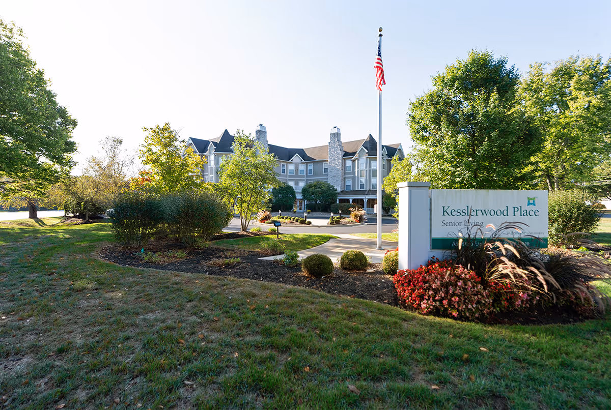 Front view of a senior living building with a sign, landscaped entrance, and flagpole.