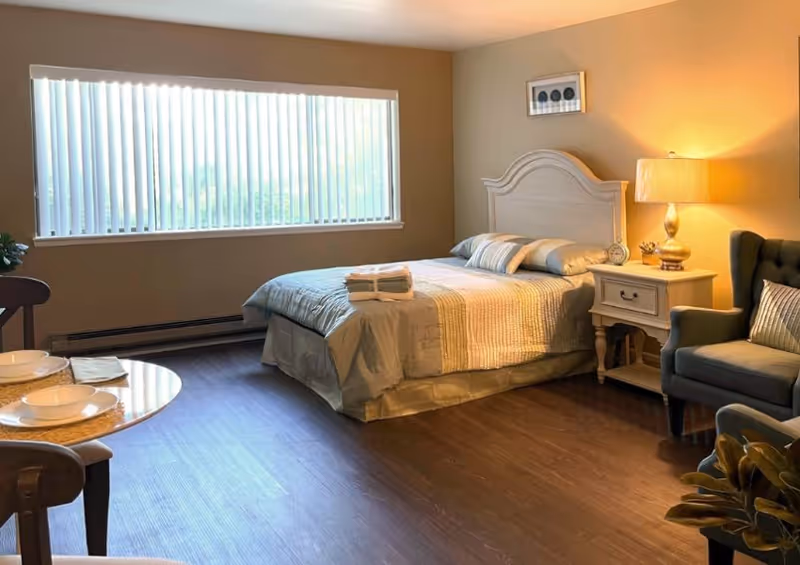 Bright bedroom with queen bed, nightstand, lamp, armchair, and small dining table.