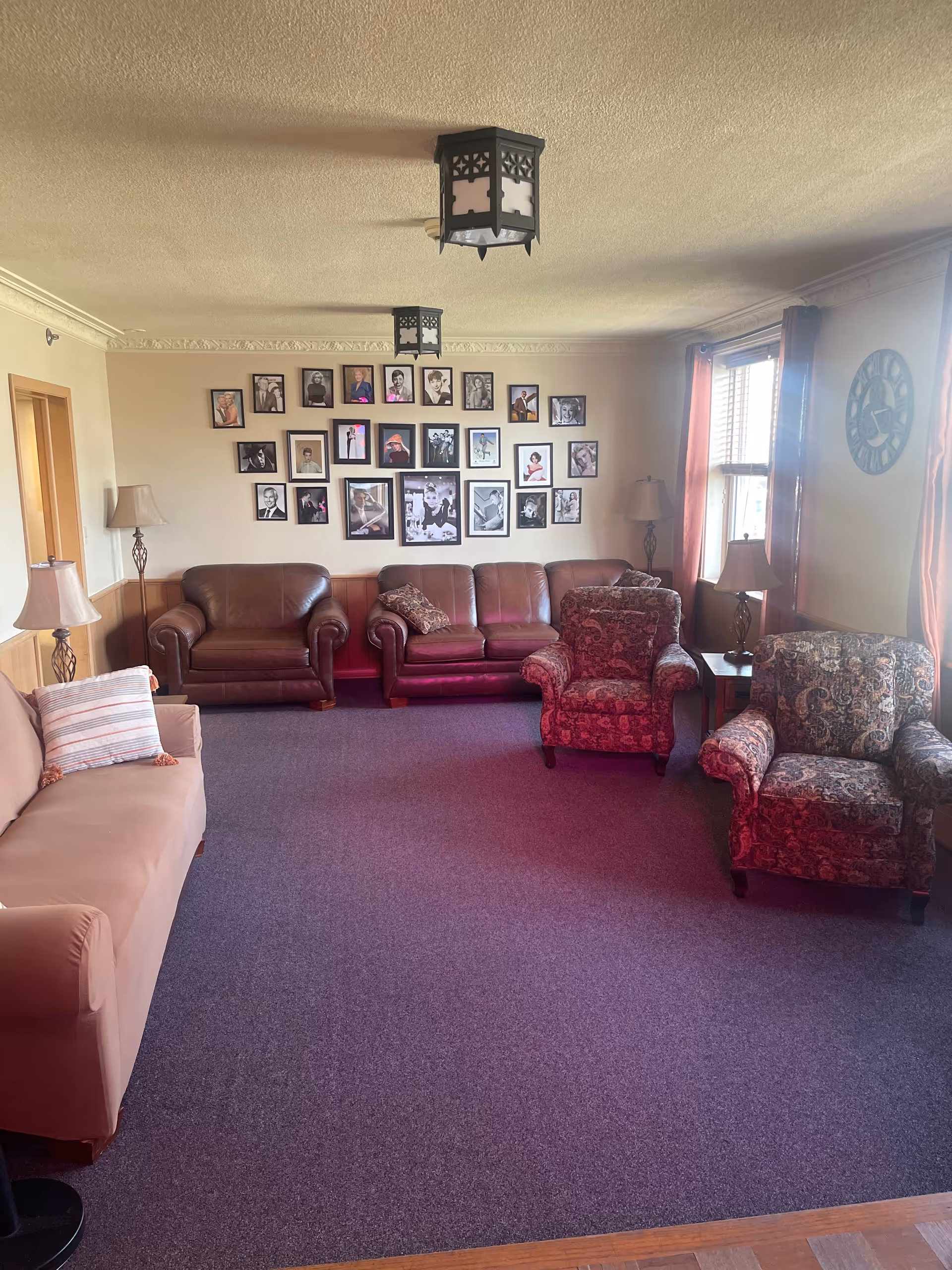 A cozy living room with multiple seating options including a beige sofa with a striped pillow, two patterned armchairs, and a brown leather loveseat and armchair. The walls are decorated with a gallery of framed black and white photographs. There are two table lamps and two floor lamps providing lighting, and a large window with red curtains letting in natural light. The ceiling has two decorative light fixtures, and a large clock is mounted on the wall near the window.