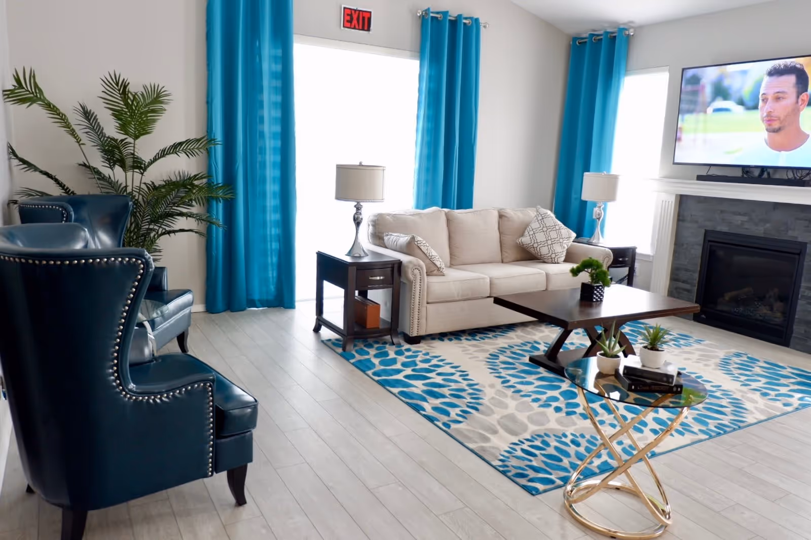 A bright living room with a beige sofa, two blue armchairs, a wooden coffee table, and a small round glass side table with plants. The room has blue curtains, a patterned blue and white rug, a fireplace with a TV mounted above it, and light-colored wooden flooring.