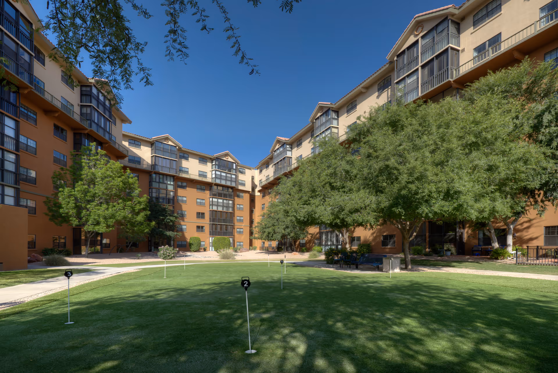 Courtyard of a multi-story senior living building with a central putting green, trees, and balconies under a clear blue sky.