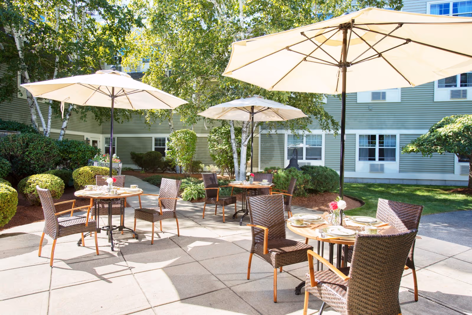 Outdoor patio area with round tables and wicker chairs under large beige umbrellas. The tables are set with plates, glasses, and napkins. The patio is surrounded by green bushes, trees, and a building with green siding and white-trimmed windows in the background.