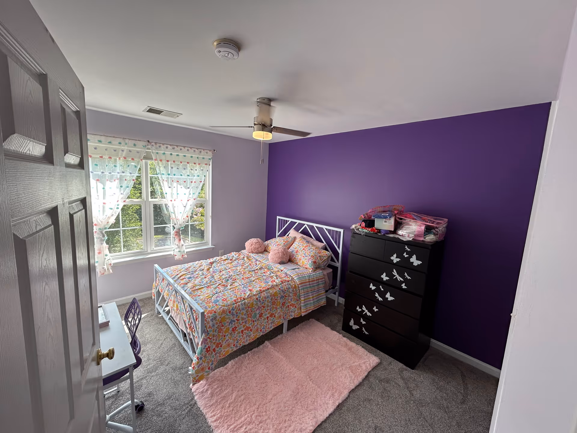 Lavender bedroom with floral quilt, pink rug, dresser, and a window with sheer curtains.