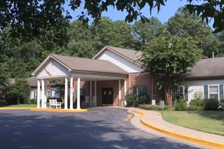 Front entrance of a brick senior living facility with a covered porte-cochère, an American flag, and a curved driveway.