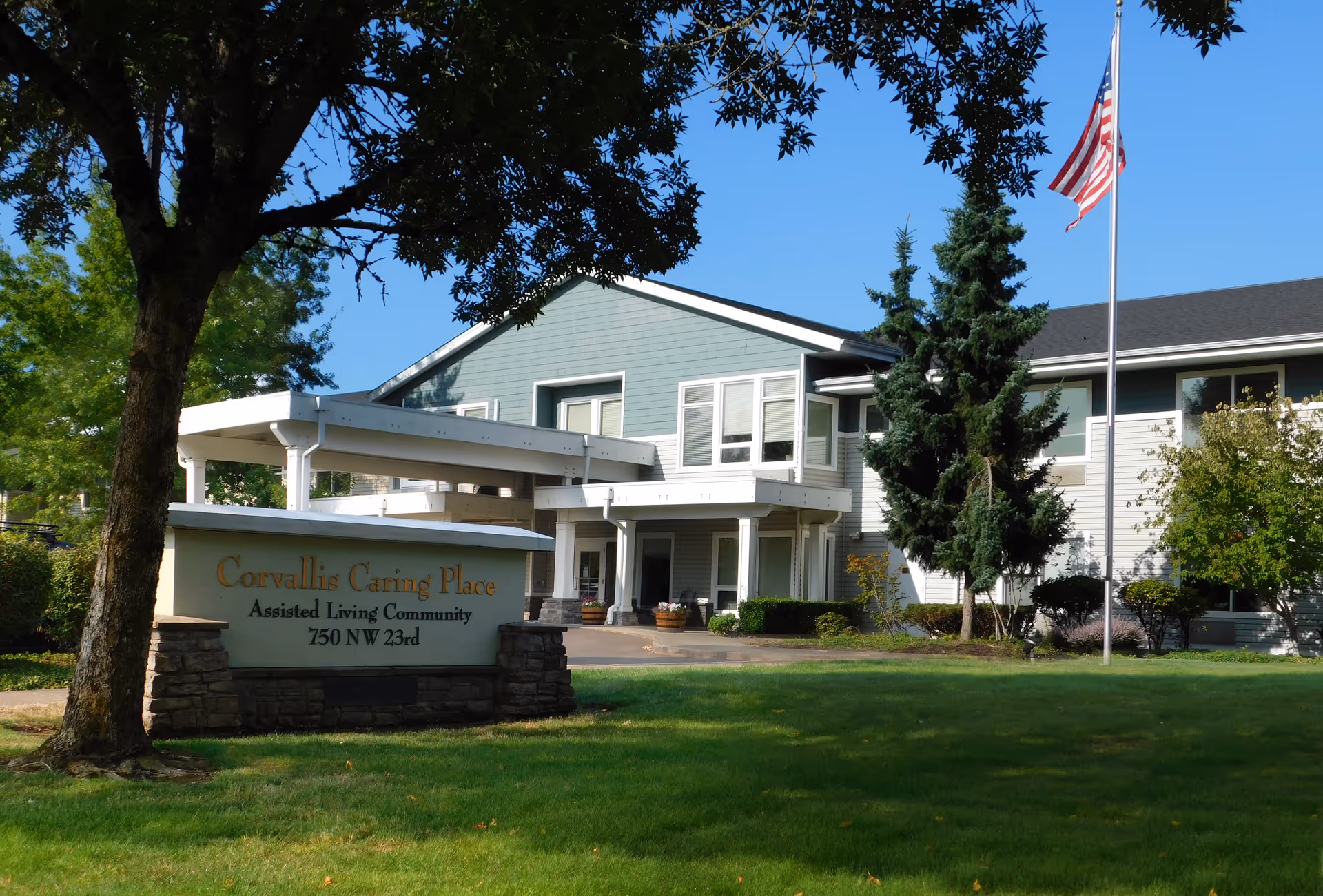 Exterior front view of Corvallis Caring Place assisted living building with entrance canopy, lawn, and American flag.