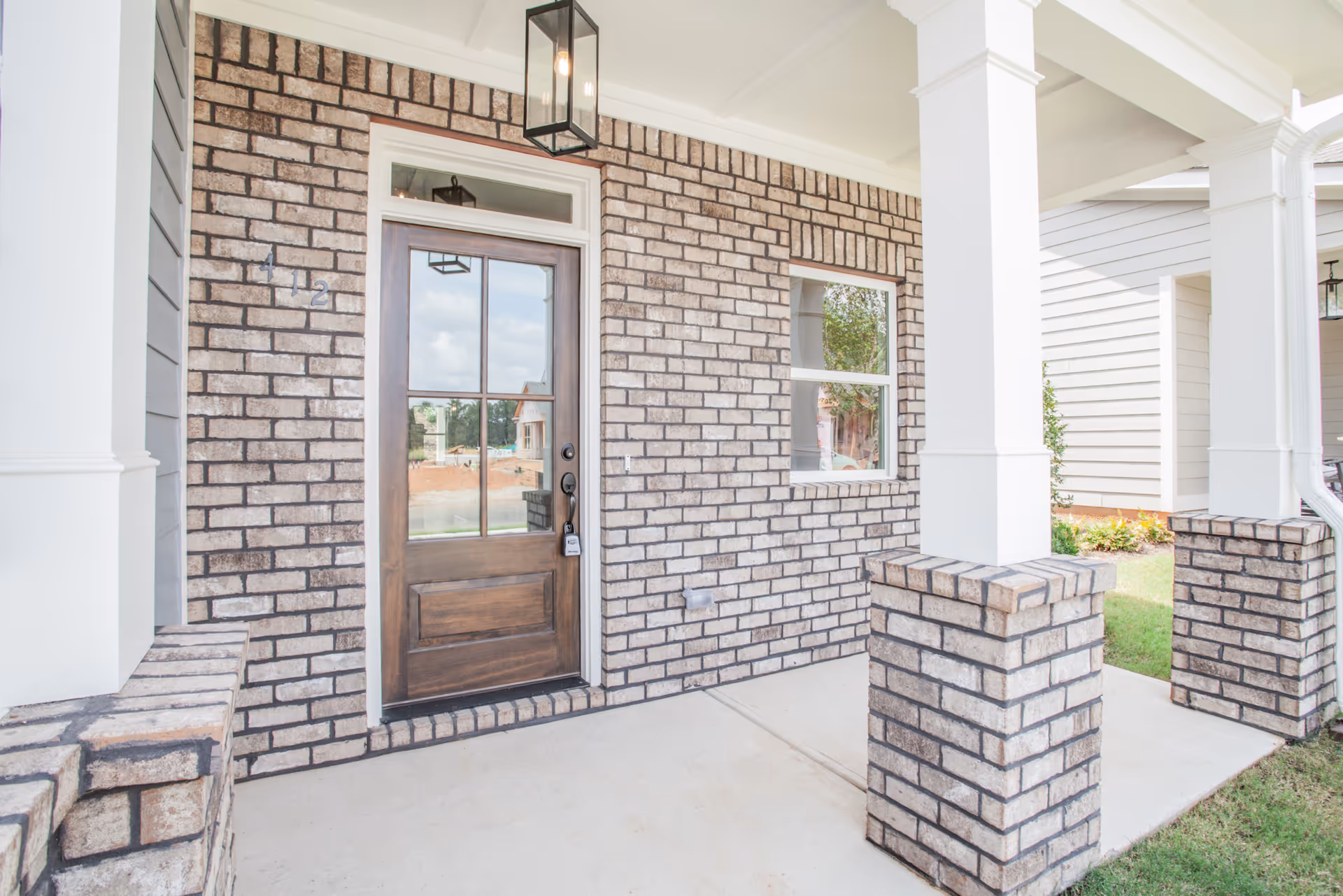 Front porch of a building with beige brick walls, a wooden door with glass panels, a window, and white columns supporting the roof. There is a hanging lantern light above the door and a concrete floor on the porch.