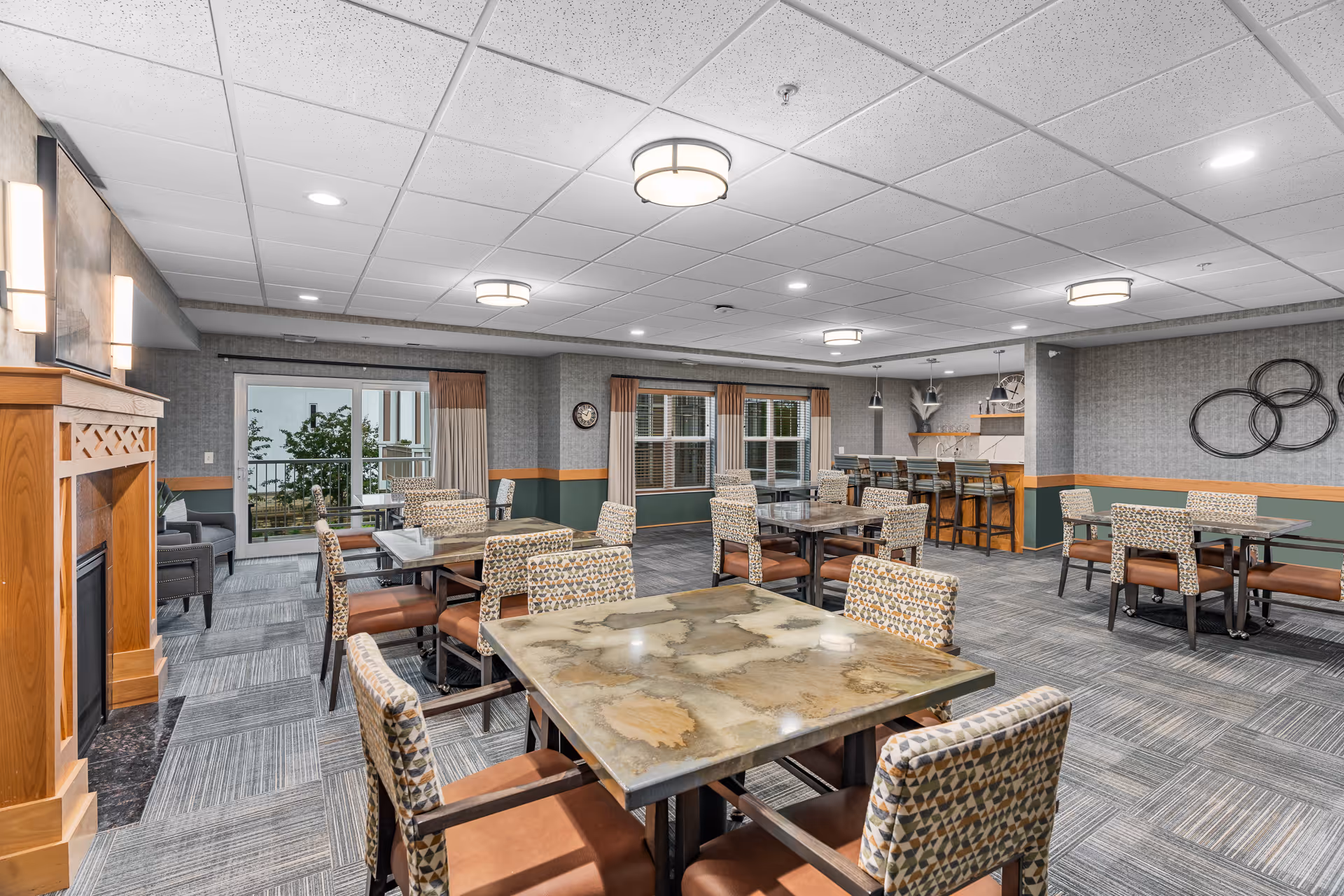 A spacious and well-lit common dining area in a senior living facility with multiple tables and chairs arranged neatly. The room features a fireplace on the left, large windows with curtains, a bar counter with stools in the background, and modern ceiling lights. The decor includes patterned chairs, a clock on the wall, and decorative wall art.