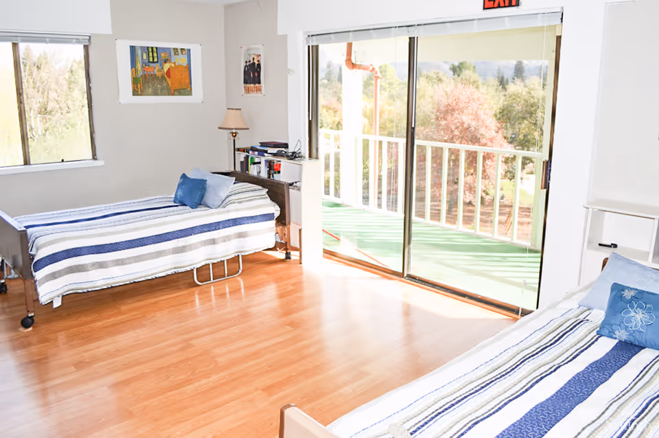 Sunlit shared bedroom with two beds, wood floors, a bedside table and a sliding glass door opening to a balcony.
