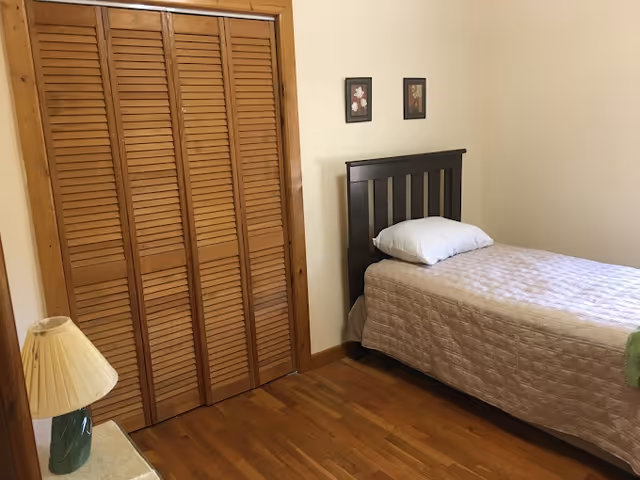 A simple bedroom with a single bed covered in a light beige quilt and a white pillow. The bed has a dark wooden headboard. There are two small framed pictures hanging on the wall above the bed. To the left, there is a wooden closet with louvered doors. A small table with a lamp featuring a green base and a beige lampshade is partially visible in the foreground. The room has wooden flooring and cream-colored walls.