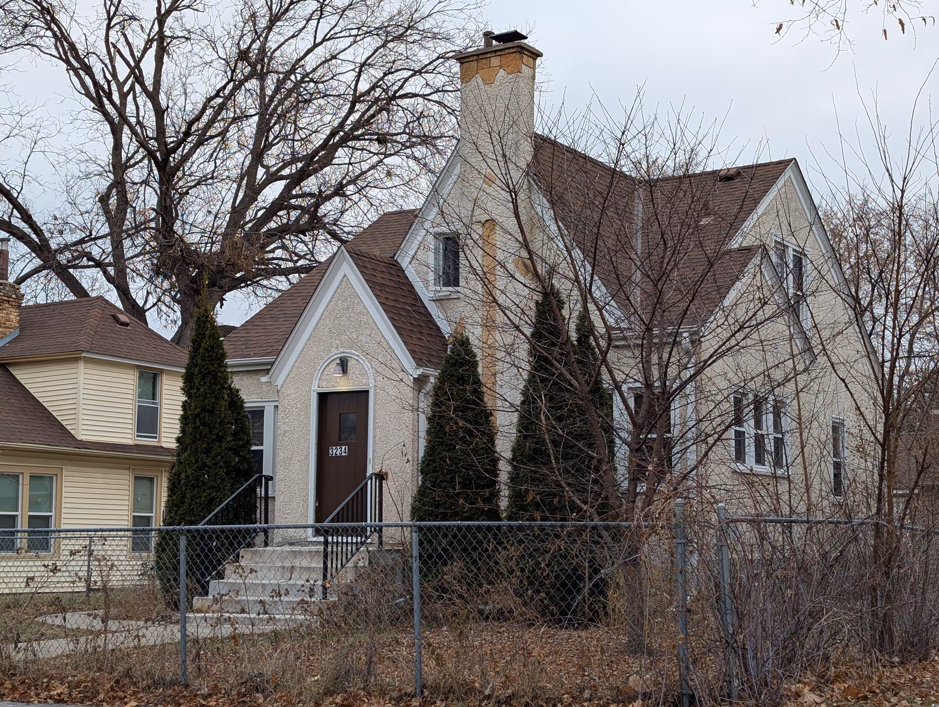 Front exterior of a two-story stucco house with a steep pitched roof, chimney, front steps and a chain-link fence, surrounded by leafless trees.