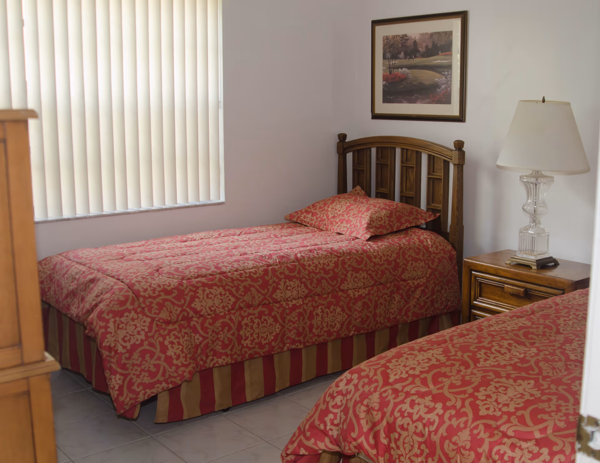 Two twin beds with red patterned bedspreads in a simple bedroom with a nightstand, lamp, and a window with vertical blinds.