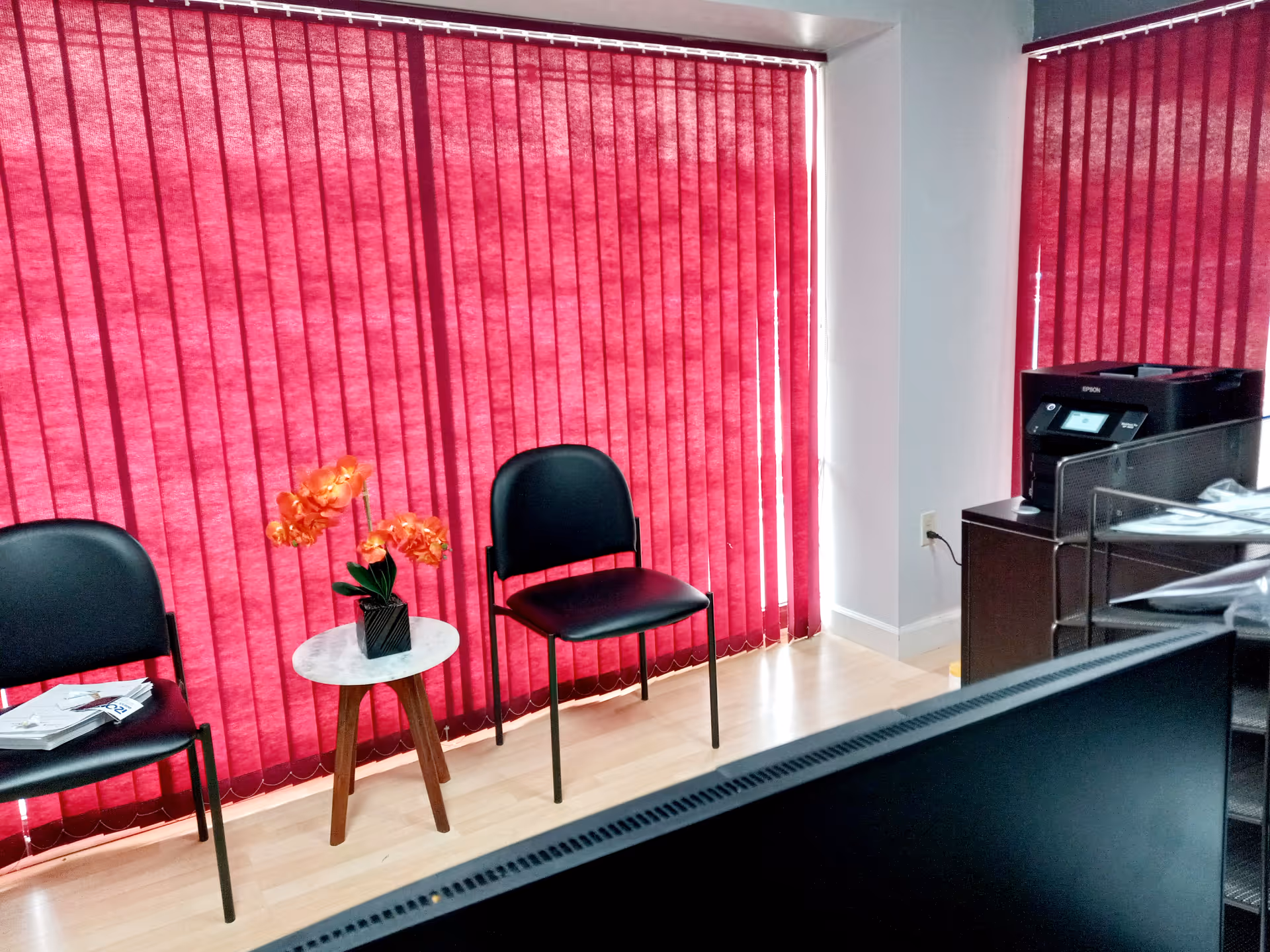 Office reception area with red vertical blinds, two chairs, and a small table with flowers.