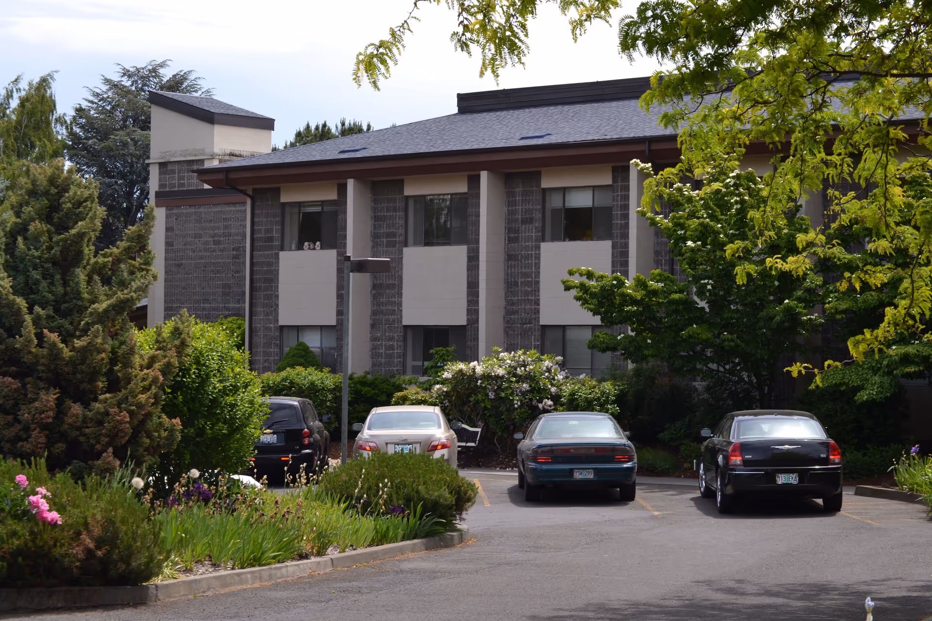 Exterior view of a two-story building with a parking lot in front. Several cars are parked, and there are trees and bushes surrounding the area, creating a green and serene environment.