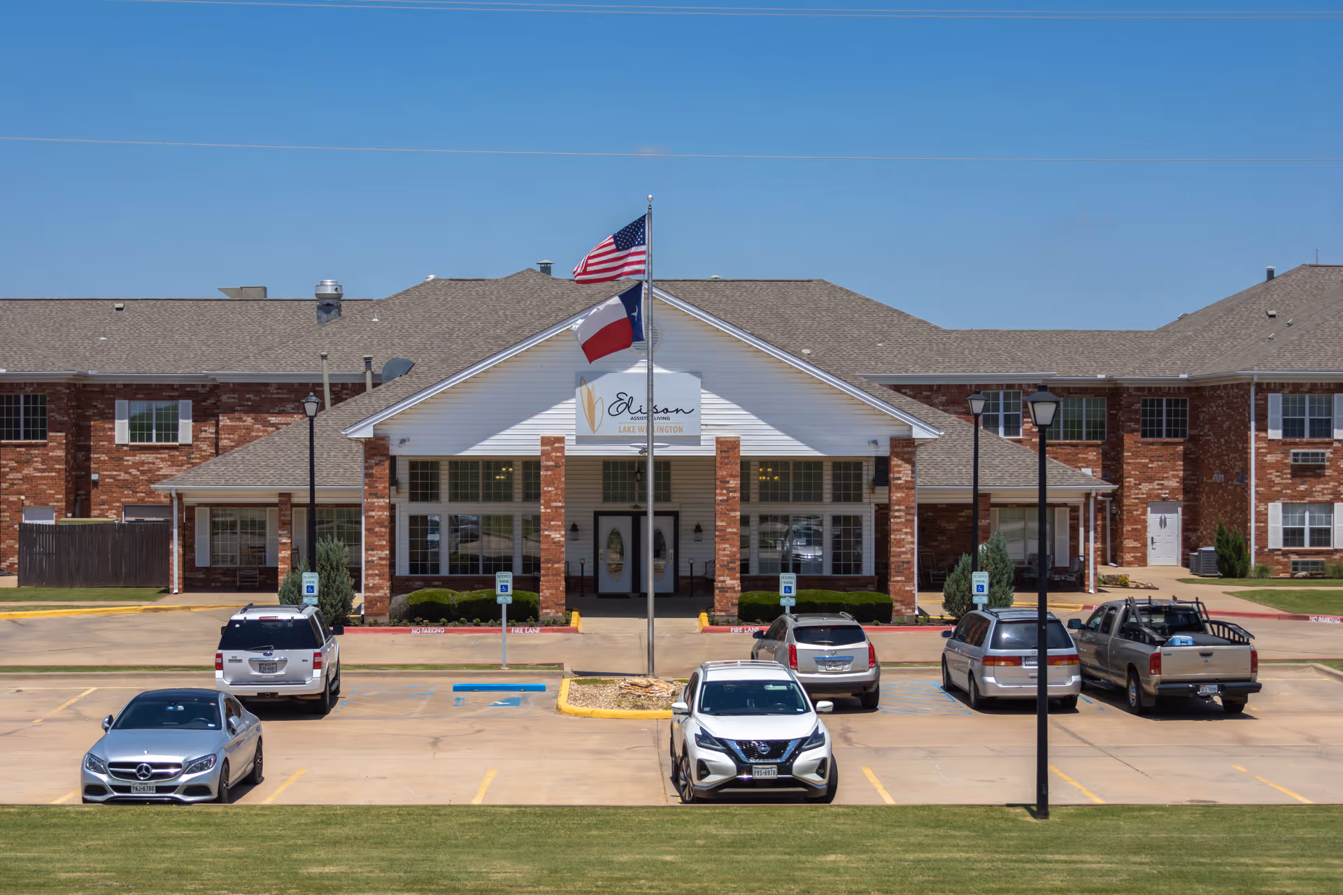 Front exterior of a brick assisted living facility with a covered entrance, flagpoles, and a parking lot with several cars.