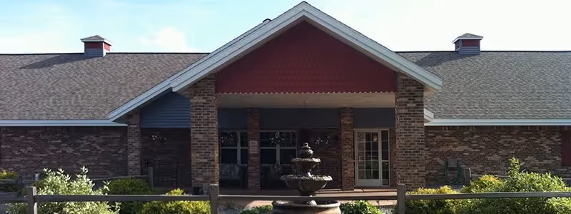 Brick single-story building with a gabled covered entrance, porch columns, and a tiered fountain in front.