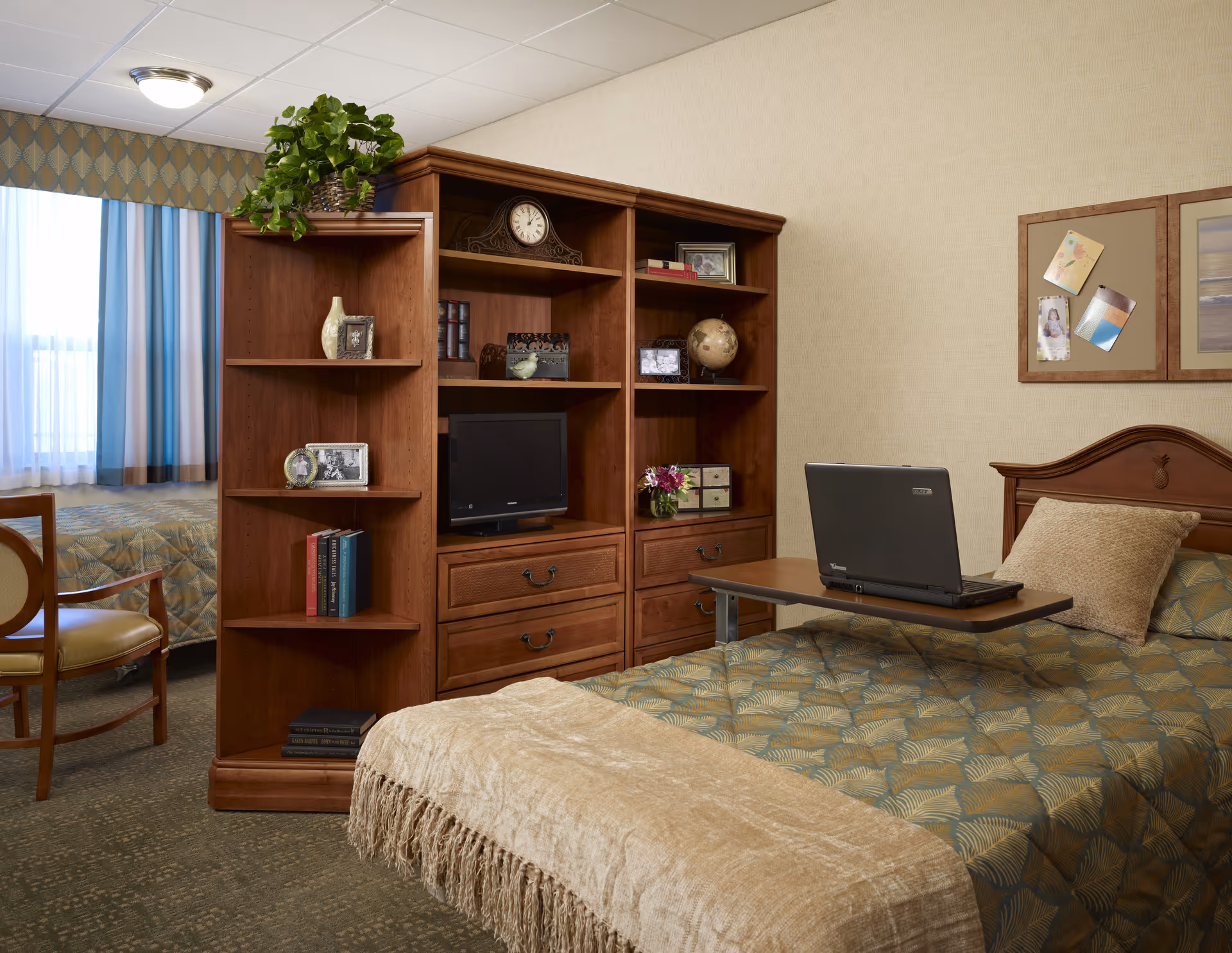 Bedroom in a senior living facility with bed, desk, bookshelf, and laptop on a tray.