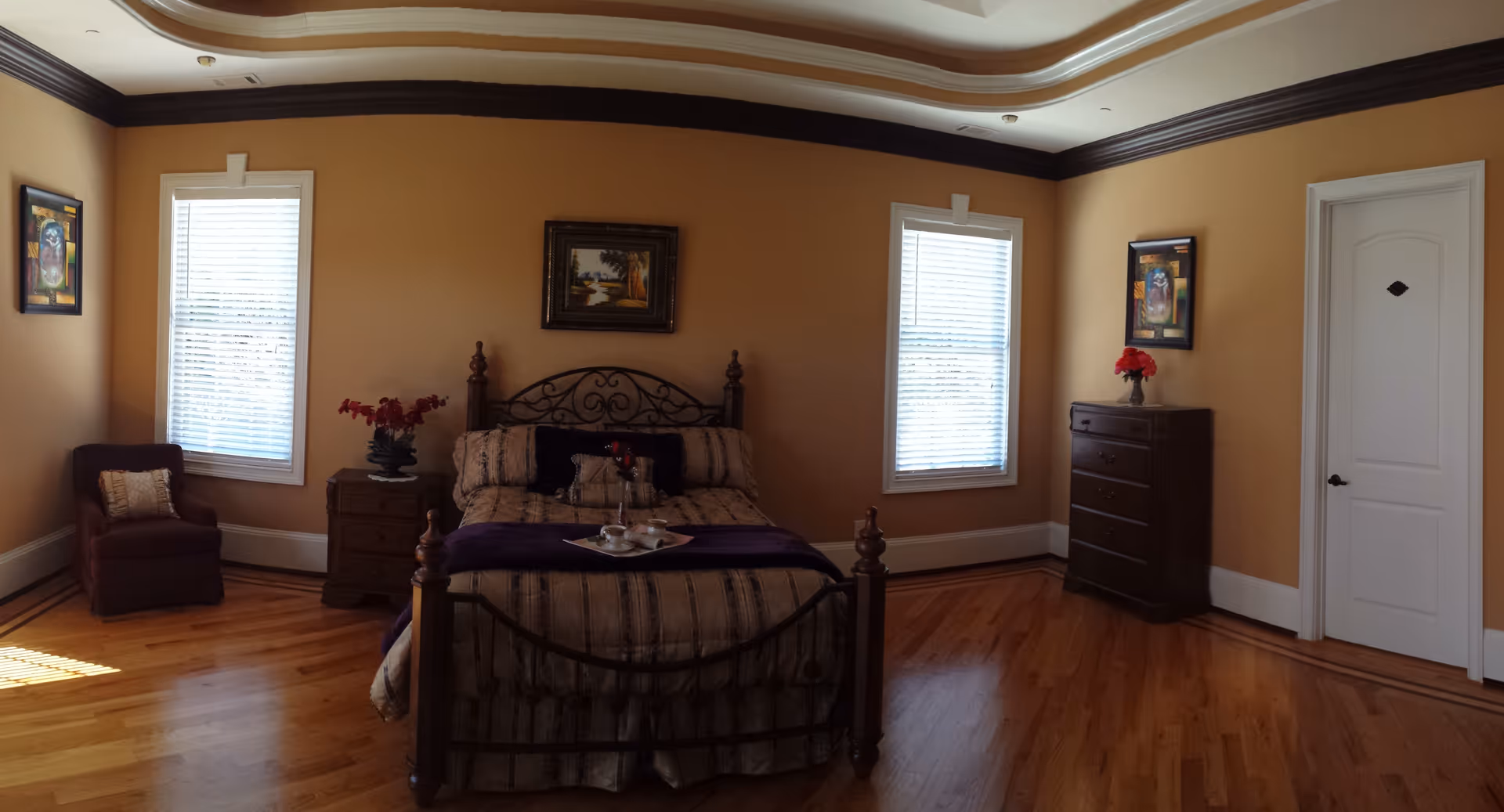 A bedroom with a wooden floor, a bed with a decorative metal headboard and footboard, beige and purple bedding, and a tray with cups on the bed. There are two windows with white blinds on either side of the bed, a wooden nightstand with a vase of red flowers to the left, a wooden dresser with a vase of red flowers to the right, a maroon armchair with a pillow in the corner, and framed artwork on the walls. The ceiling has a decorative molding and recessed lighting.