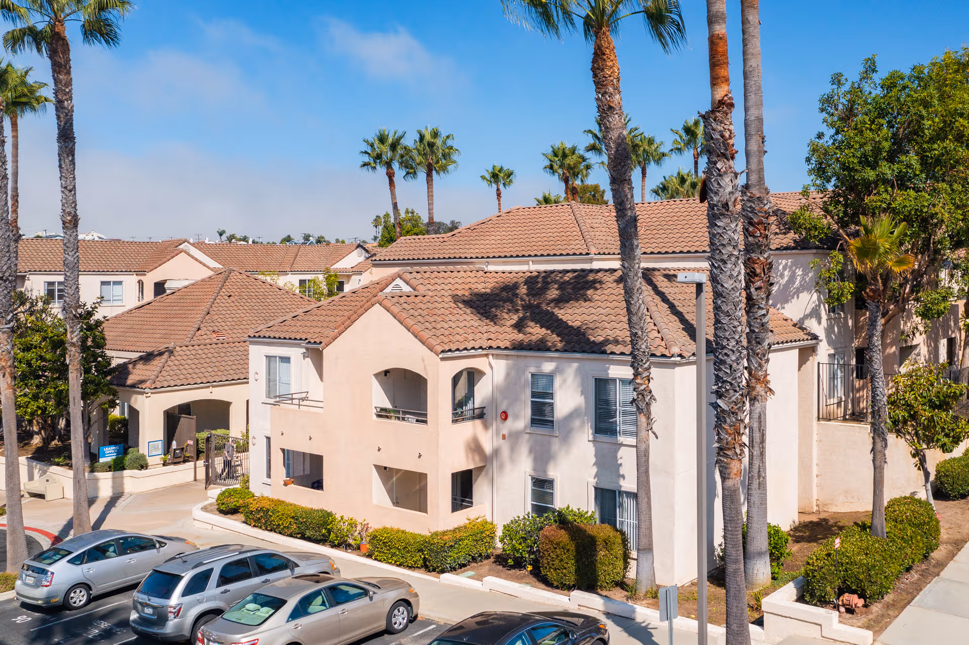 Exterior view of a beige senior housing complex with red-tiled roofs, palm trees, and parked cars.