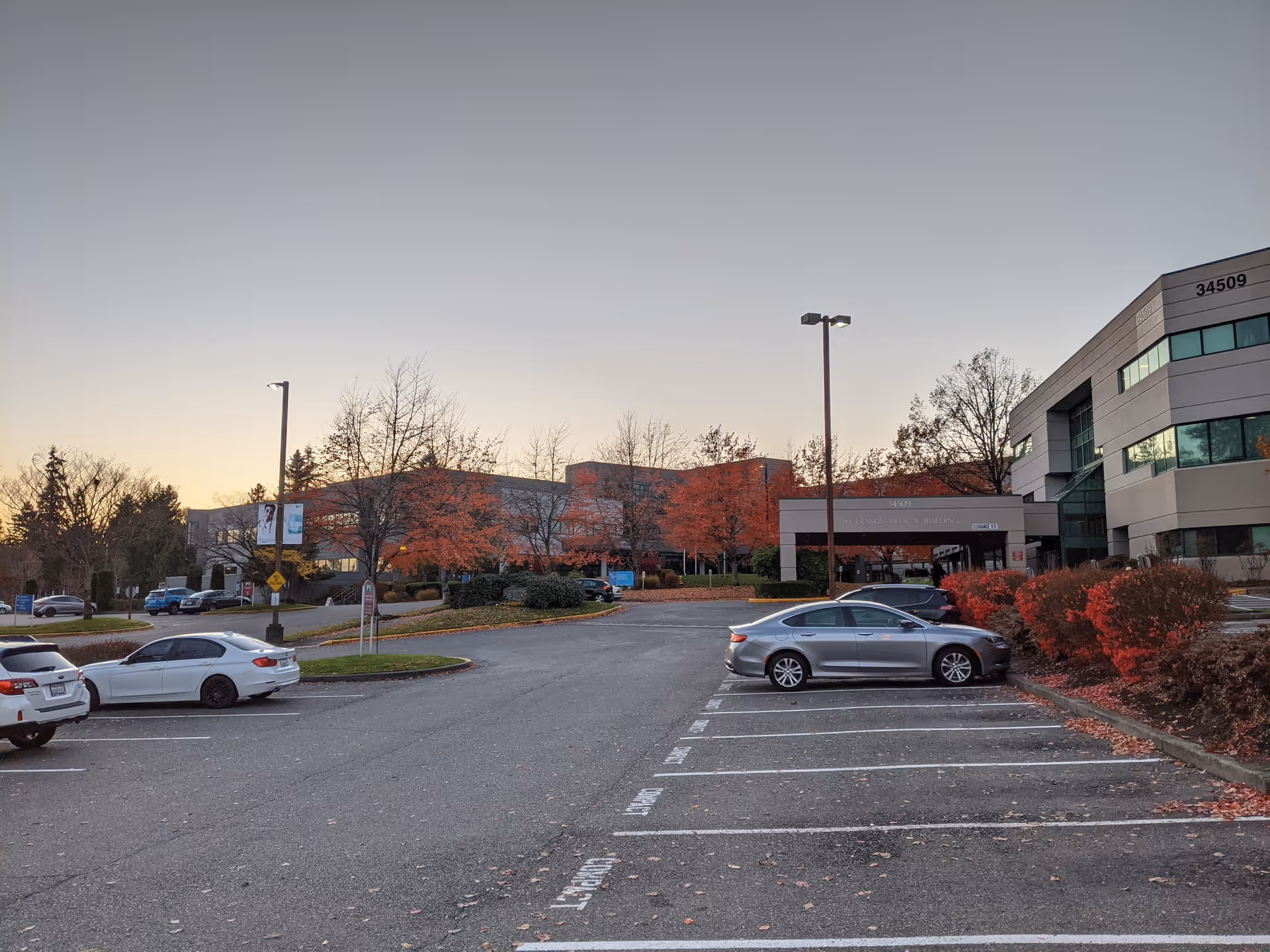 Parking lot with cars in front of a modern hospital-like building amid autumn trees.