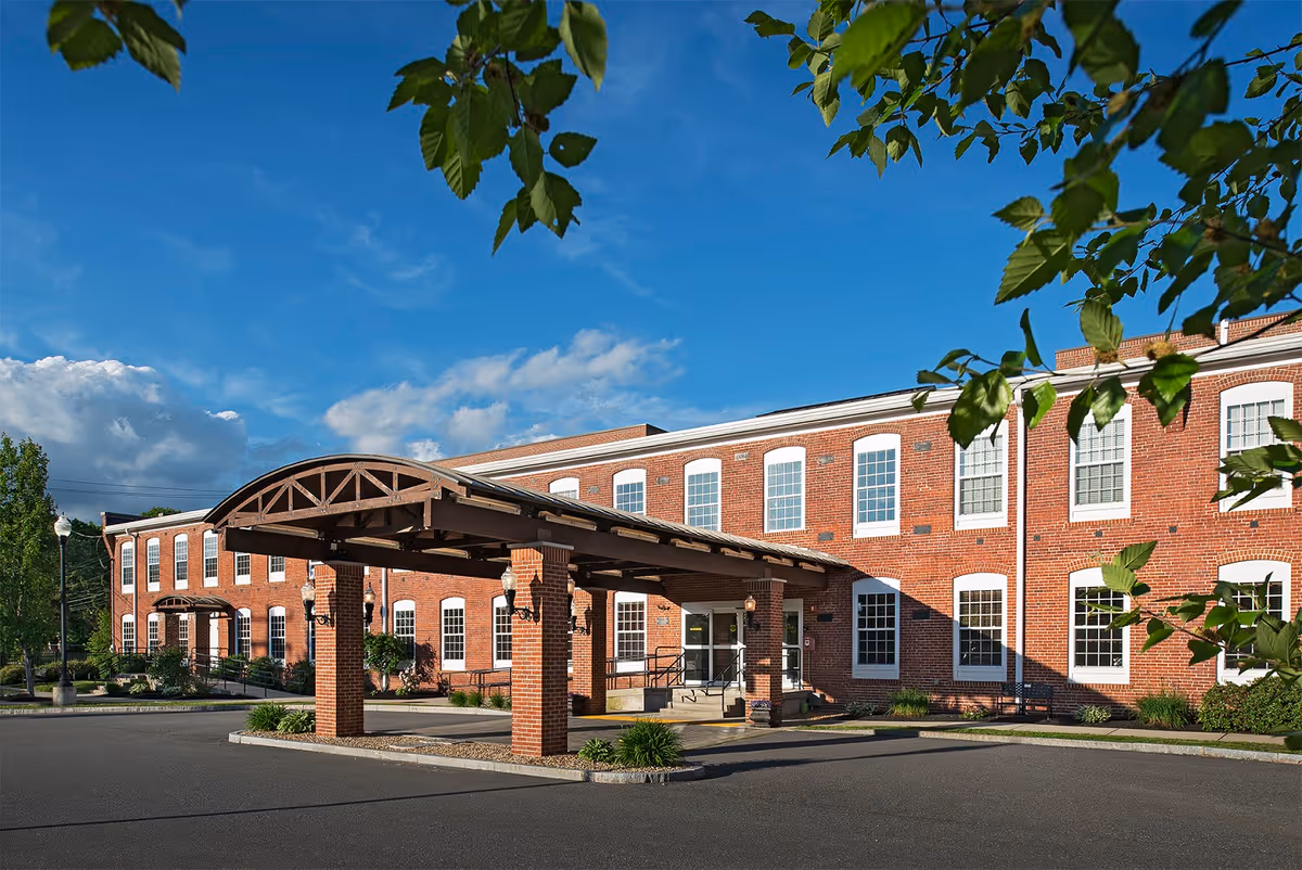 Exterior view of a two-story brick building with large windows and a covered entrance supported by brick pillars. The sky is clear with some clouds, and green leaves from nearby trees frame the top of the image.
