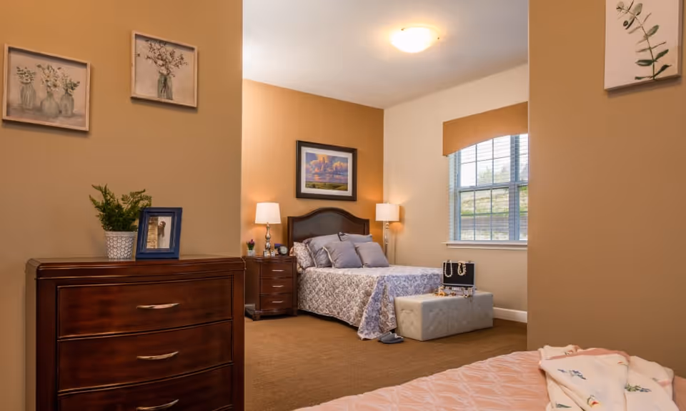 A cozy bedroom in a senior living facility with a bed covered in a patterned quilt, two bedside tables with lamps, a window with blinds, a cushioned bench at the foot of the bed displaying jewelry, a wooden dresser with a plant and framed photo, and floral artwork on the walls.