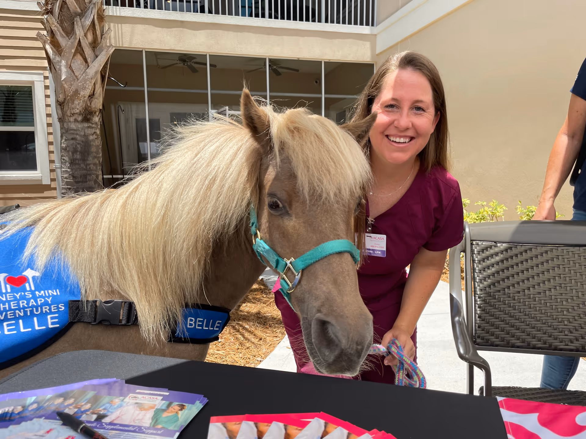 Smiling caregiver in scrubs with a small therapy pony outdoors at ACASA SeniorCare.