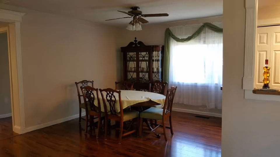 A dining room with a wooden dining table covered by a light-colored tablecloth and six matching wooden chairs with green cushions. There is a wooden china cabinet against the wall, a ceiling fan with lights above the table, and a large window with sheer white curtains and green drapes. The floor is hardwood, and there is a small counter with a decorative amber glass bottle on the right side.