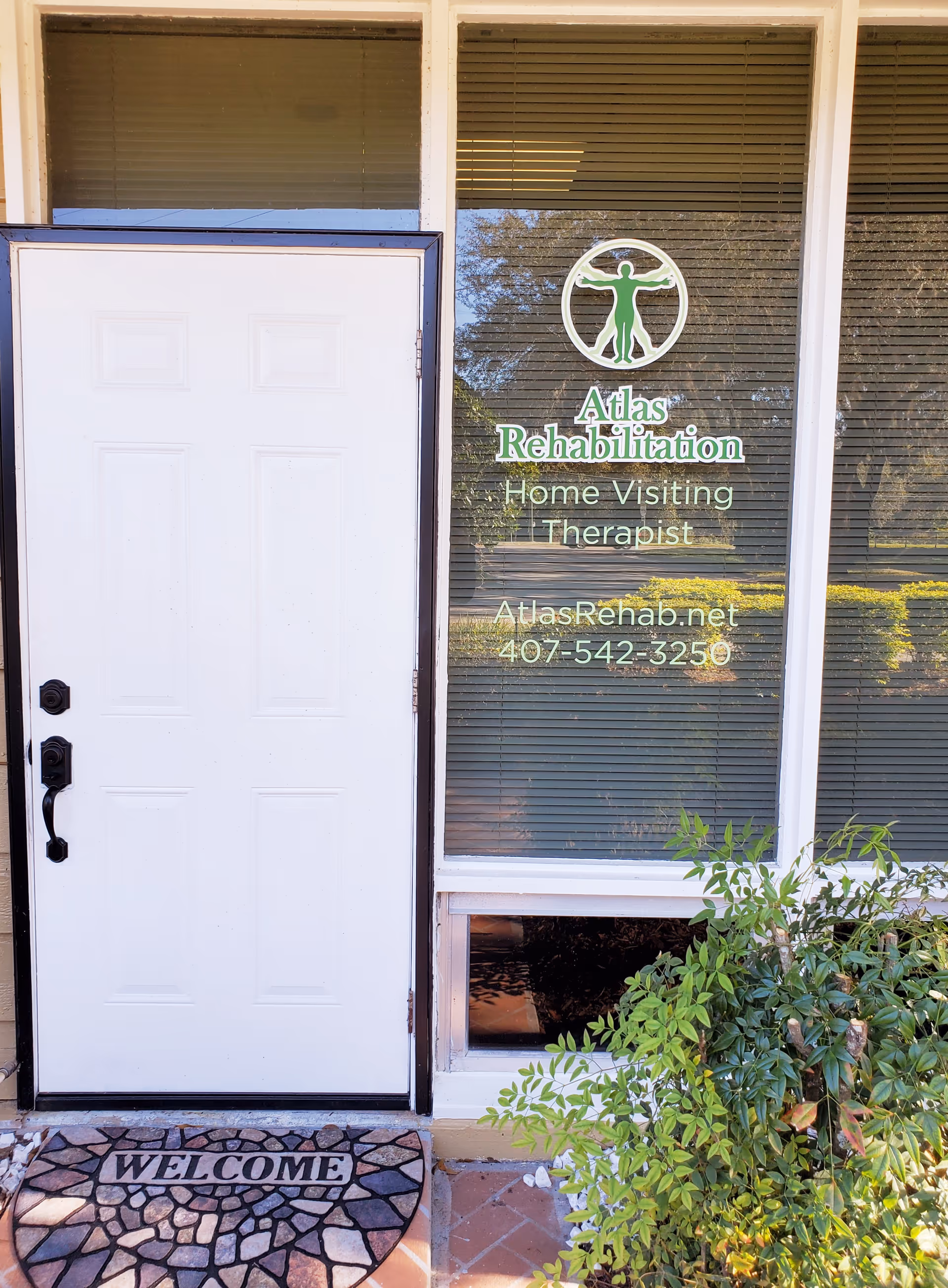 White entry door beside a window with Atlas Rehabilitation signage and a welcome mat.