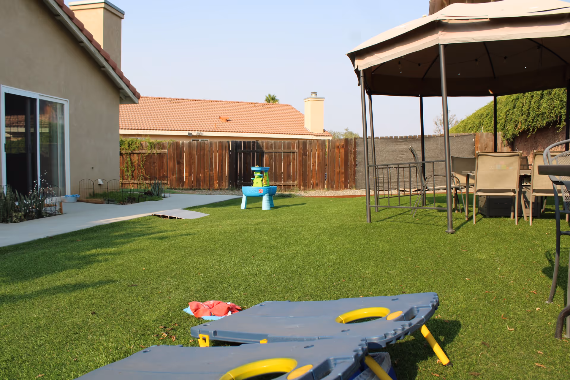 Outdoor area of a senior care facility with green grass, a gazebo with chairs and a table, a small blue water play table, and a cornhole game set up on the lawn. There is a beige building with a sliding glass door on the left and a wooden fence in the background.